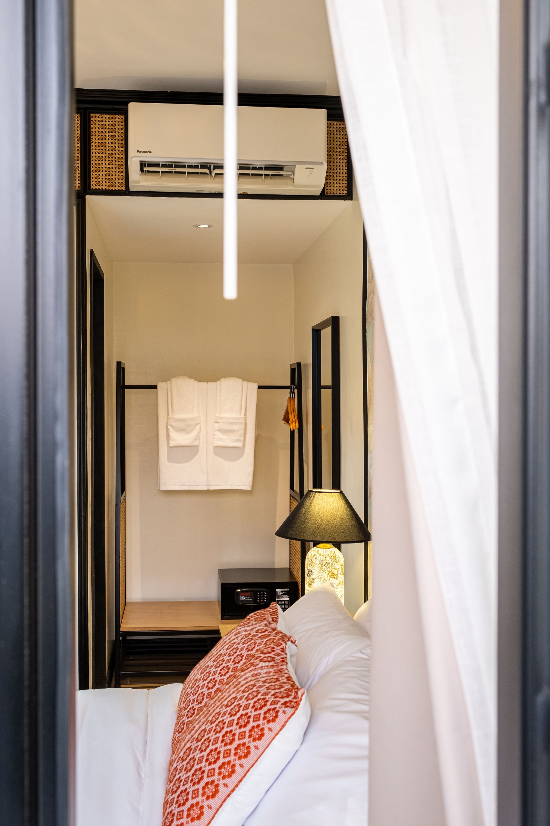 A view into a hotel room through a doorway, showing a bed with an orange patterned pillow, a lamp, and hanging robes.