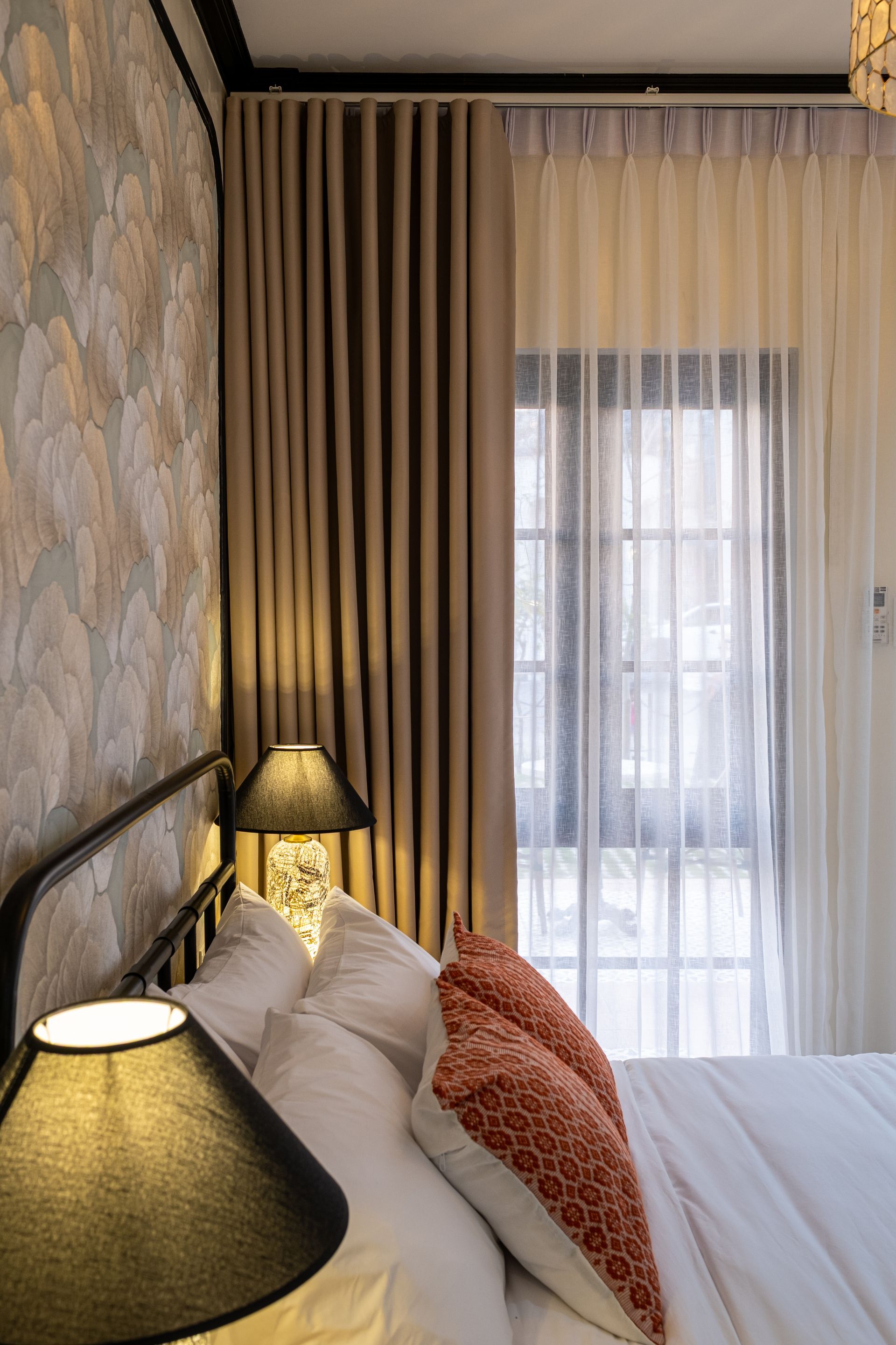 A bedroom with patterned wallpaper, a bed with white and orange pillows, two lamps, and beige and sheer white curtains.