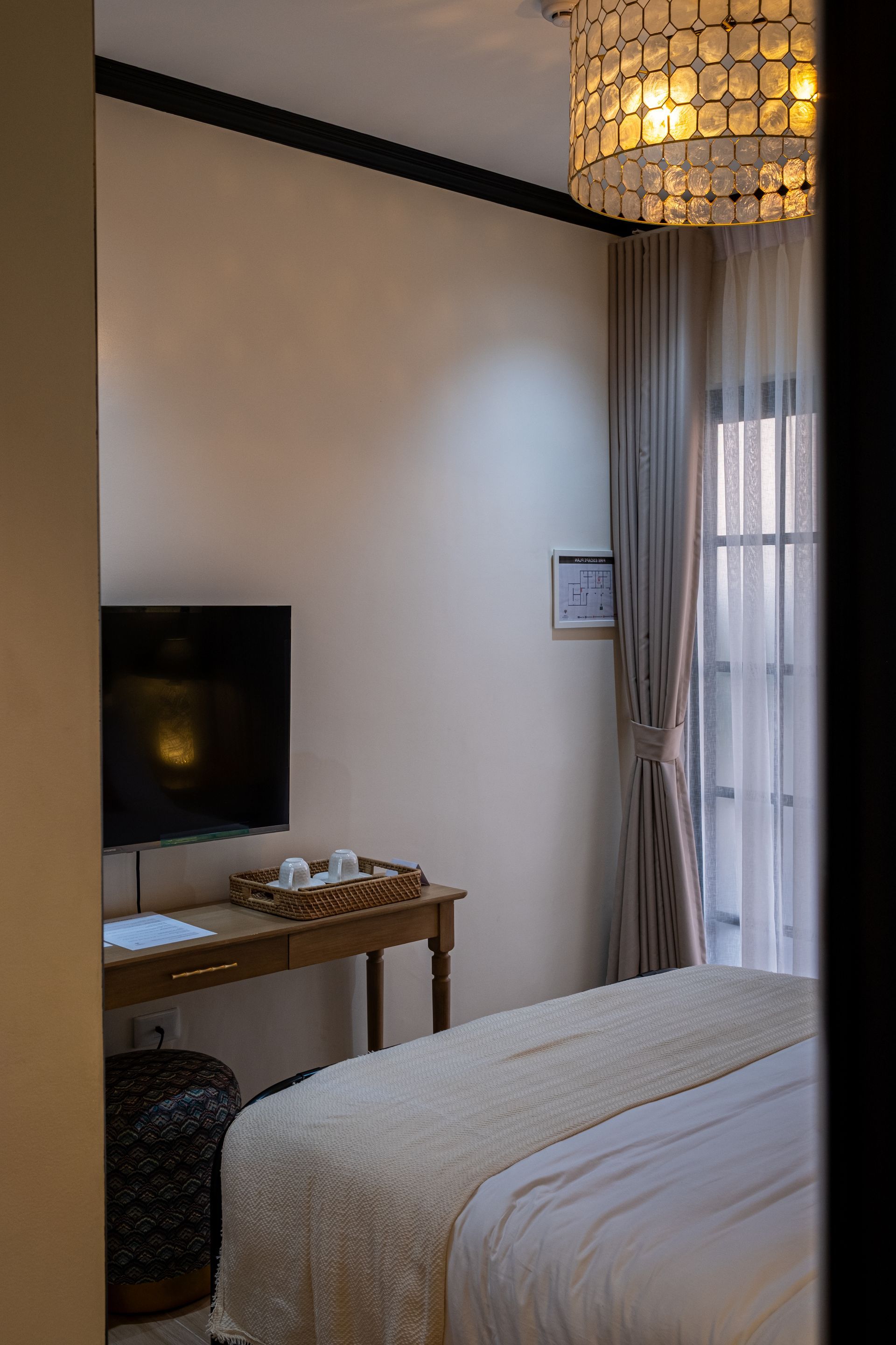 A bedroom with a bed, a decorative chandelier, a wall-mounted TV, and a small desk in a neutral-toned room.
