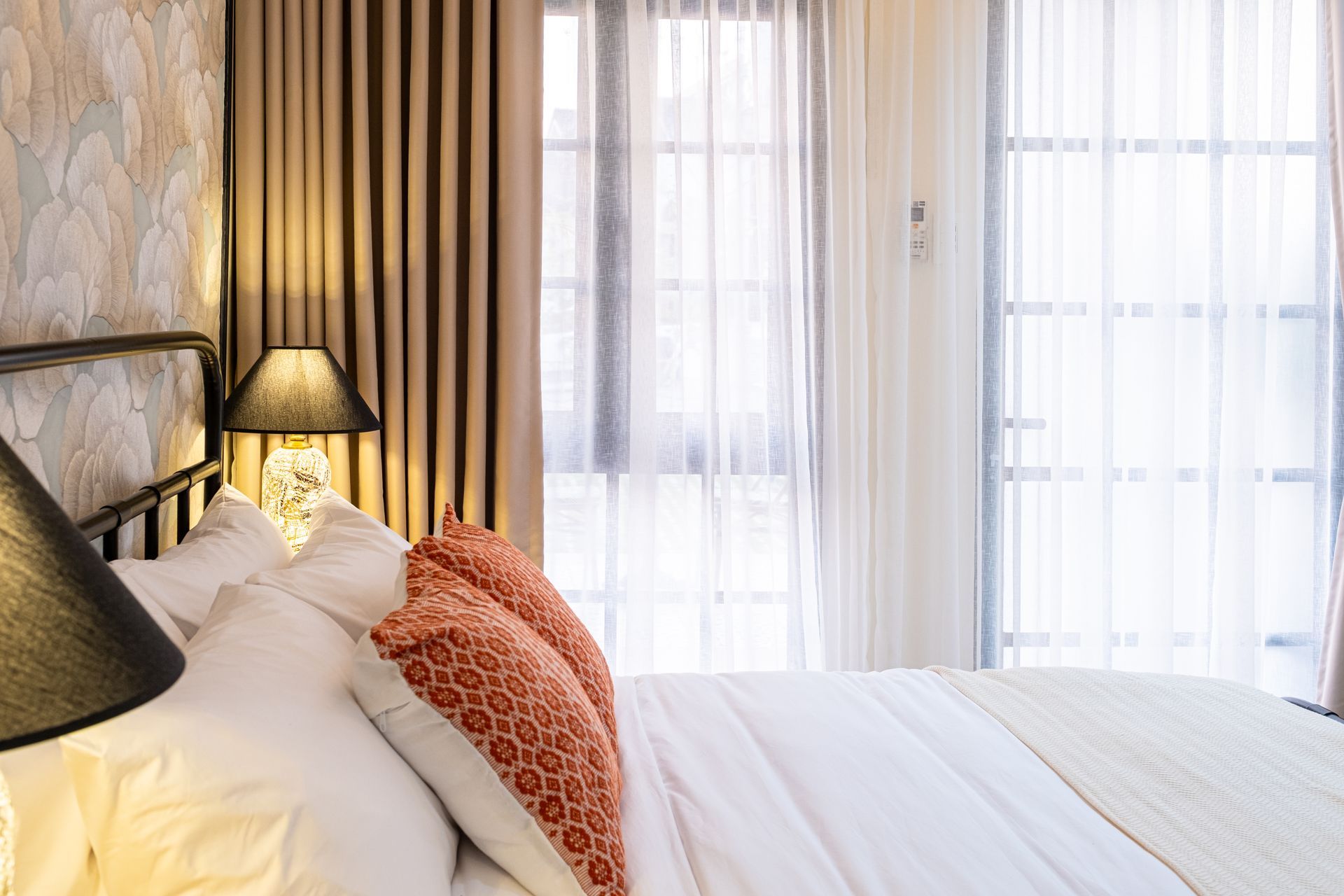 A bedroom with a bed featuring white pillows, orange patterned cushions, a patterned wallpaper, and sheer curtains.