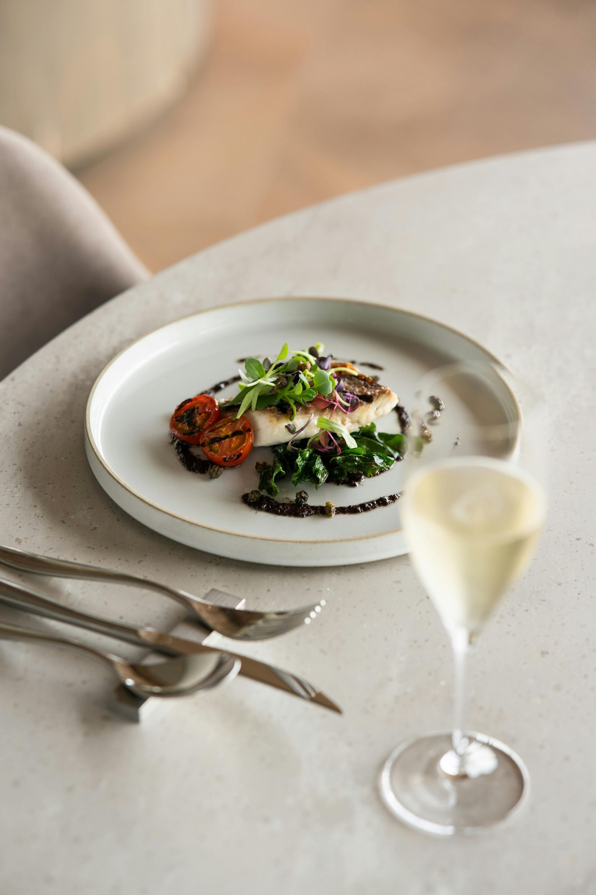 A white plate with pan-seared fish and greens, accompanied by a glass of sparkling wine on a neutral-toned table.