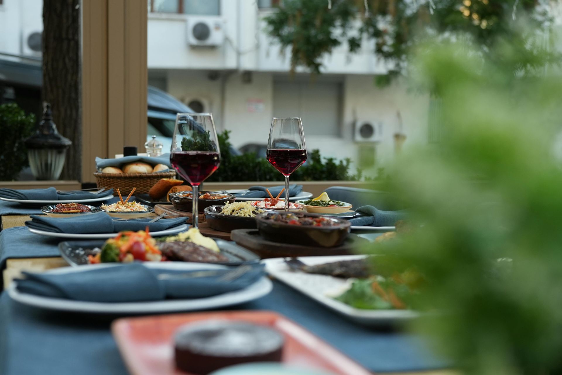 A table set for dining outdoors with two glasses of red wine, various dishes, and blue napkins on a wooden surface.