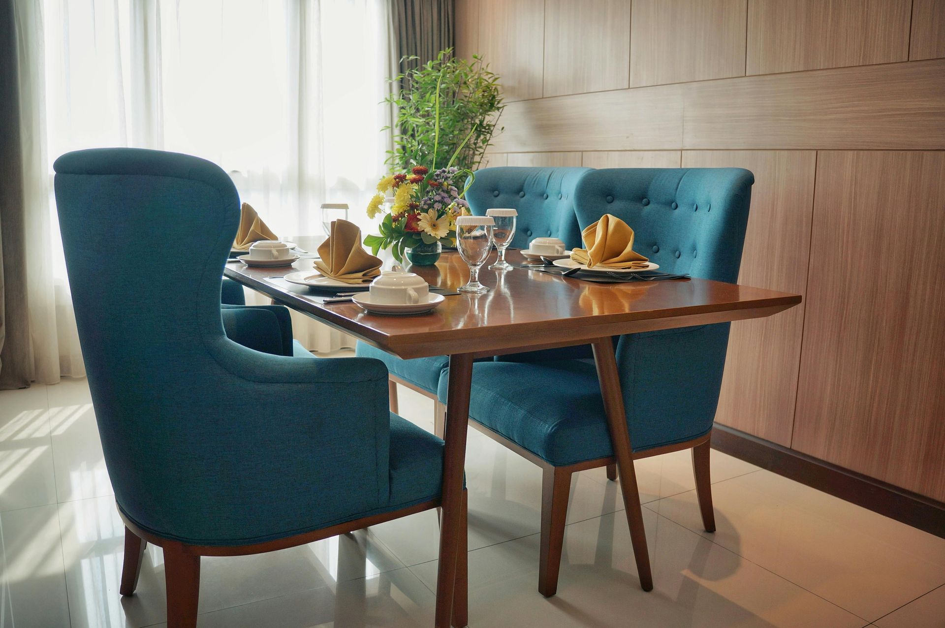 A wooden dining table with four blue tufted chairs, set with napkins and glasses, next to a window with sheer curtains.