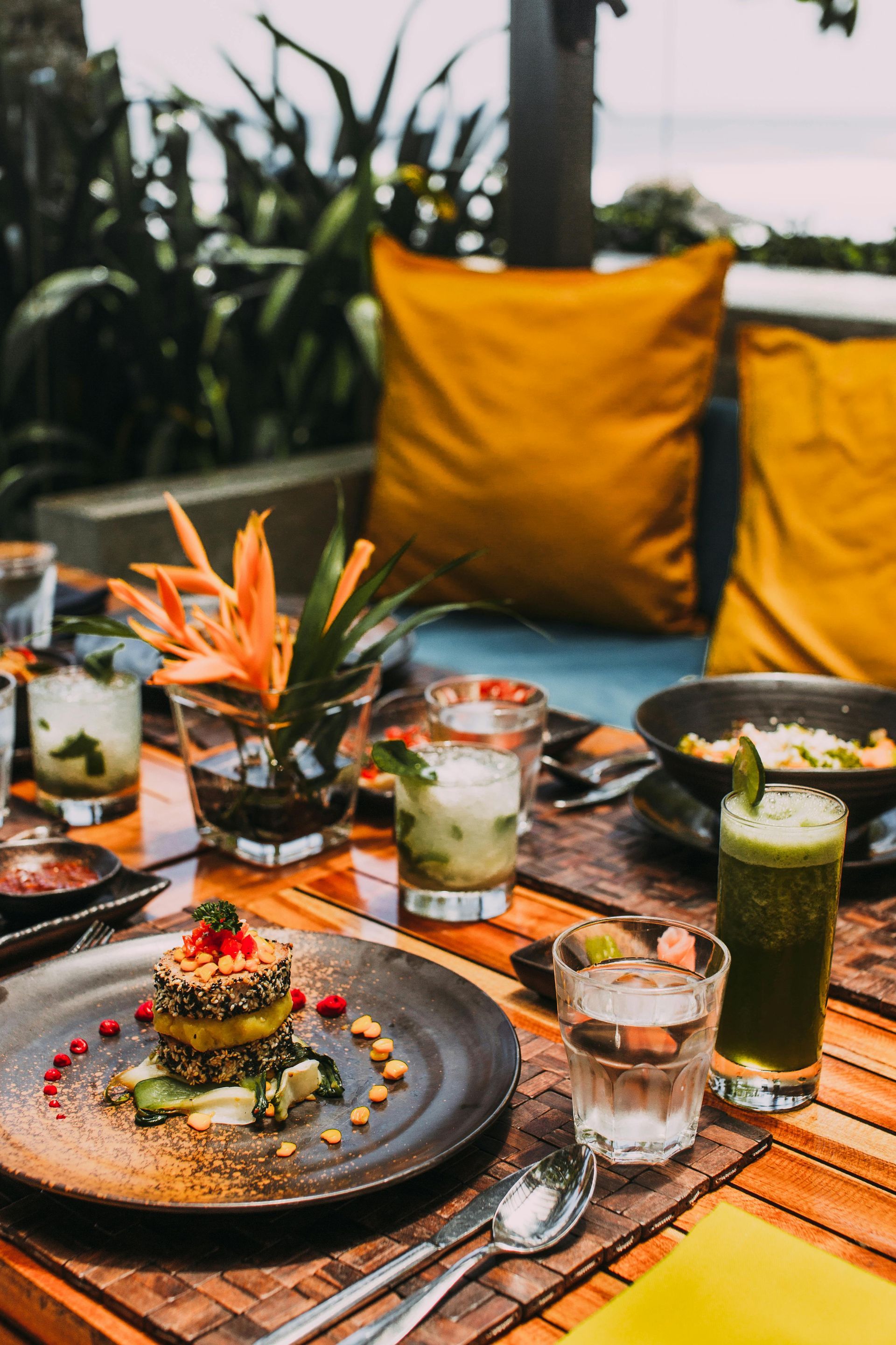 A plated meal, a green drink, and water glasses on a wooden table, with yellow cushions and greenery in the background.