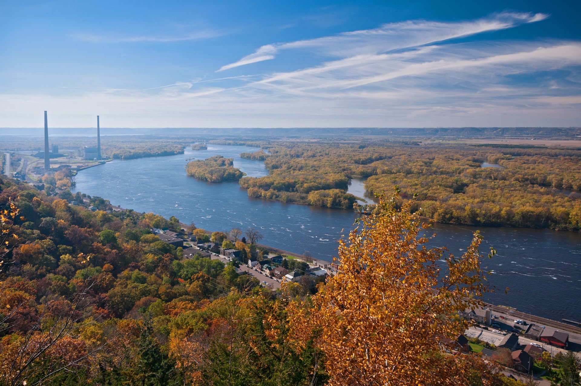 River flowing through autumn trees, under a blue sky, with smokestacks visible in the distance.