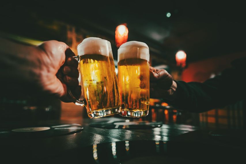 Two beer mugs clinking together in a dimly lit bar, golden beer with white foam, hands raised in a toast.