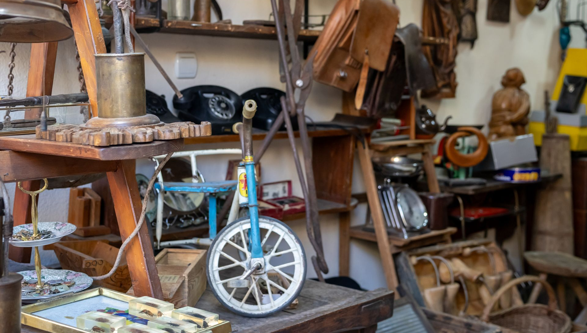 A blue vintage-style child's tricycle sits on a display stand amidst various antique objects in a shop.