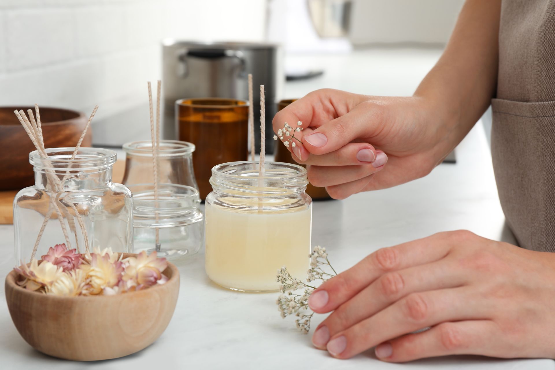 A person crafting a candle, placing dried flowers into a small jar filled with melted wax on a white surface.