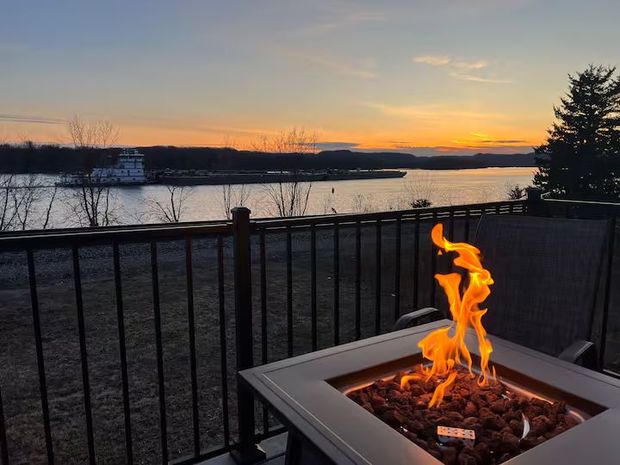 Sunset view over a river with a lit fire pit on a patio. A boat is on the water.
