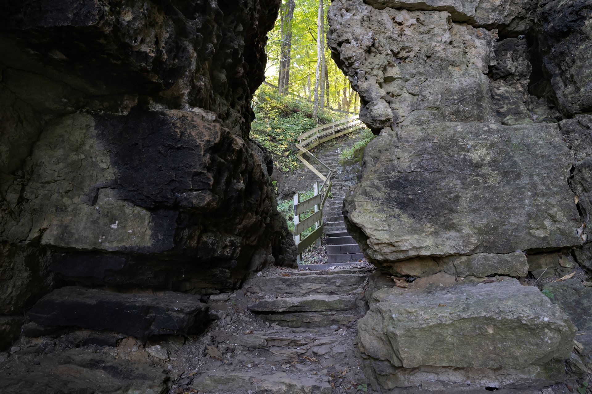 Rocky cave opening framing a wooden stairway leading up into a sunlit forest.