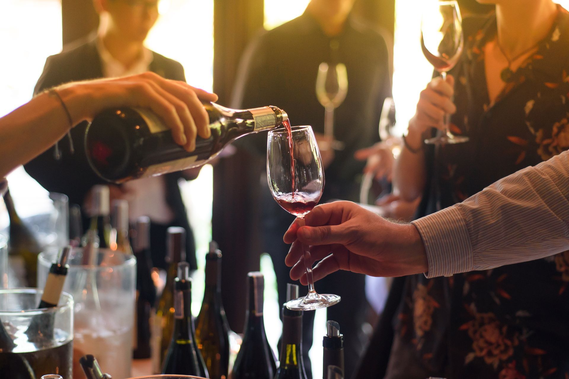 Pouring red wine into a glass at a wine tasting event, with people and bottles in the background.