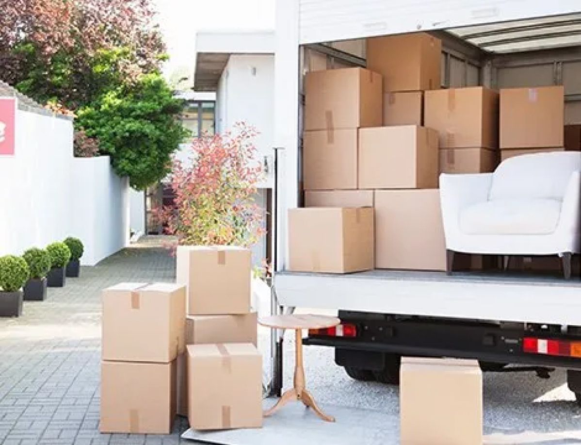 Two movers in blue coveralls load cardboard boxes into a moving truck in front of a house.
