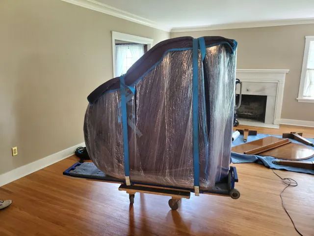 Two people loading a wooden piano onto a moving truck using a ramp, outdoors.
