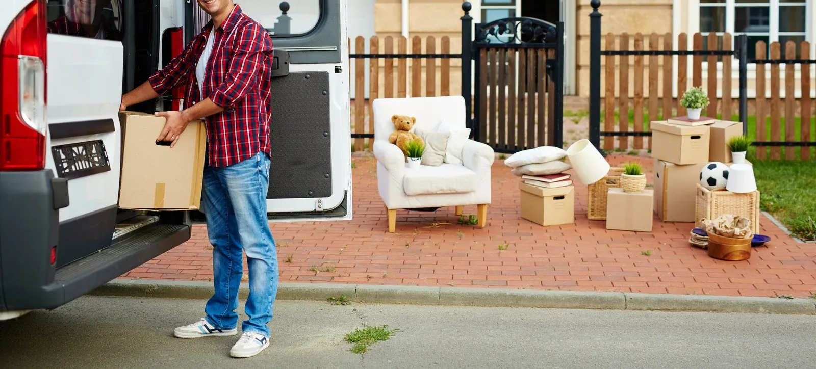 Man loading cardboard box from van onto a brick patio with furniture and boxes, in front of a house.