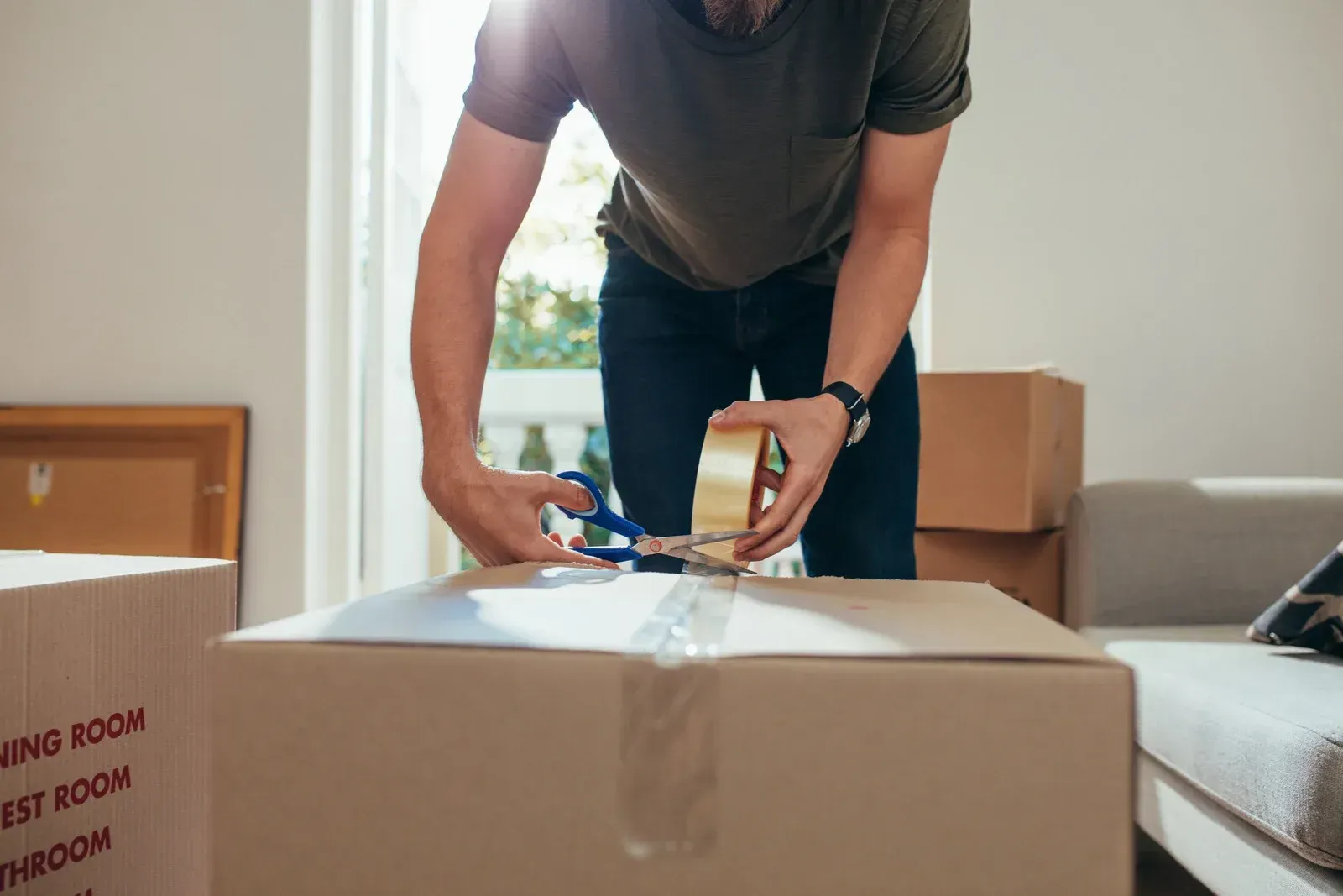 Man taping a cardboard box in a room with other boxes.