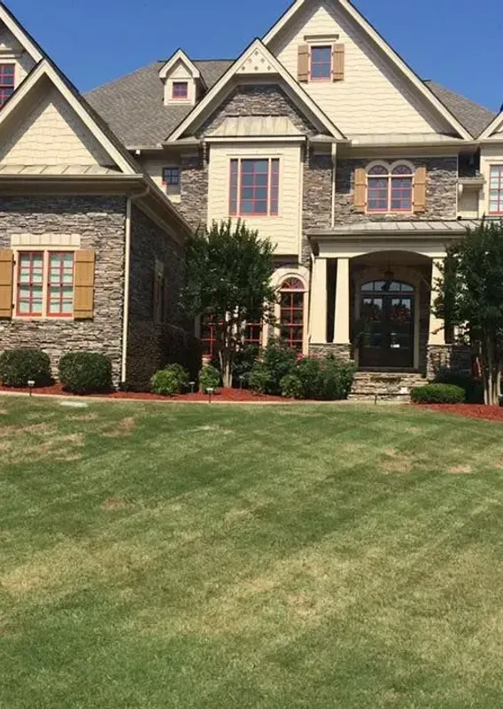Large tan stone house with a manicured green lawn under a clear blue sky
