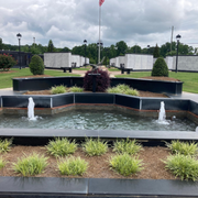 Tiered black fountain with water jets, surrounded by shrubs and flowers in a landscaped park under cloudy skies