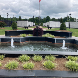Tiered black fountain with water jets, surrounded by shrubs and flowers in a landscaped park under cloudy skies