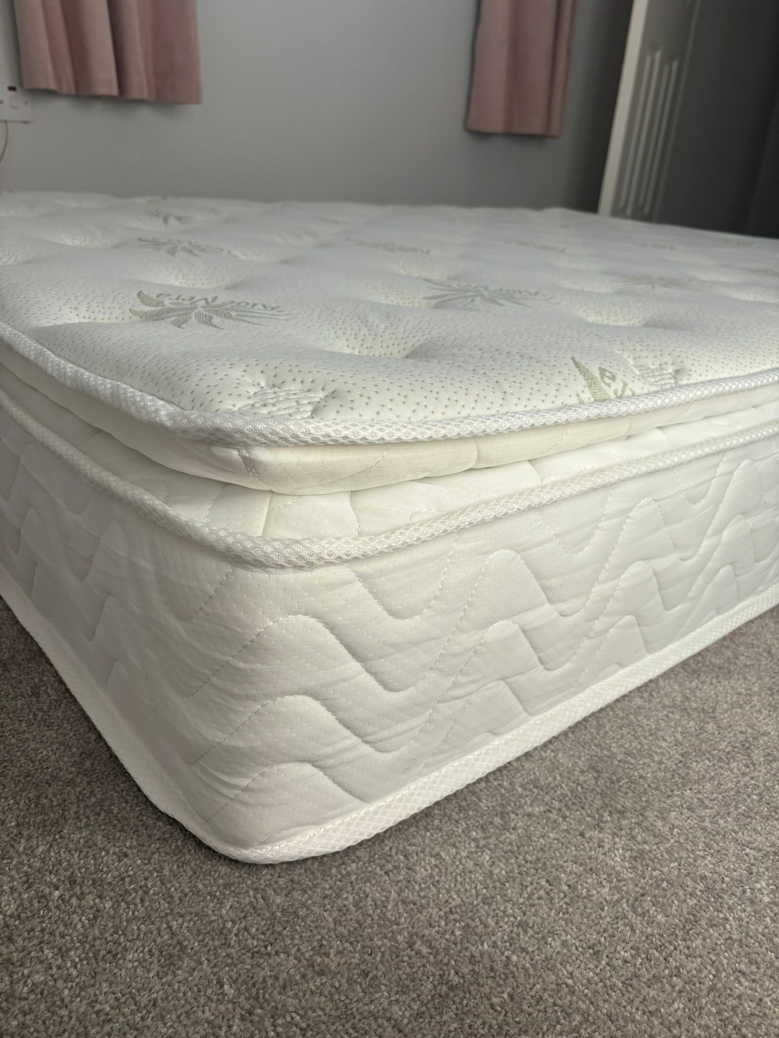 White mattress with a patterned topper on a carpeted bedroom floor near pink curtains.