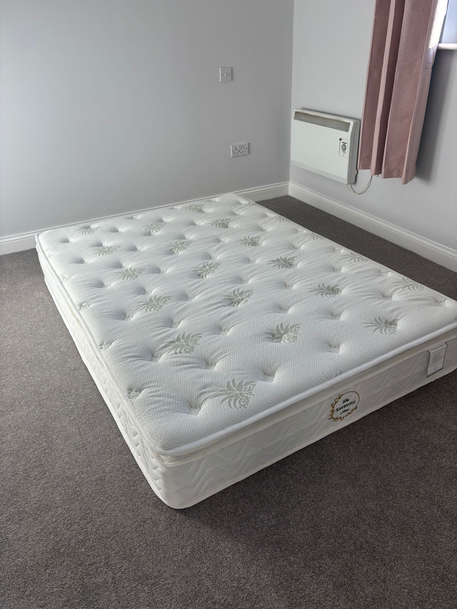 Mattress on carpeted floor in a sparsely furnished room, with gray walls and a window curtain.
