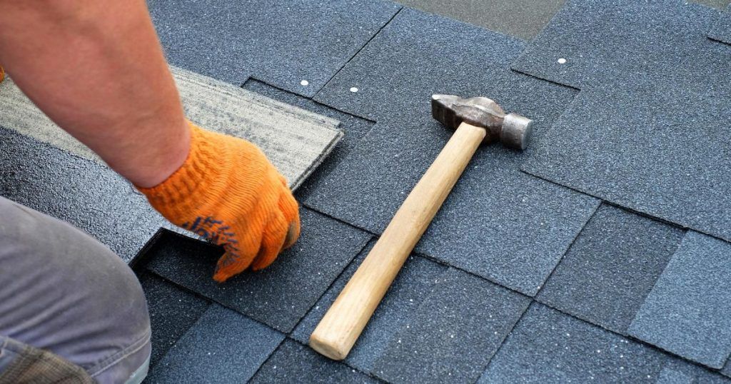 Worker fitting roof shingles with a hammer and trowel on a gray shingle roof