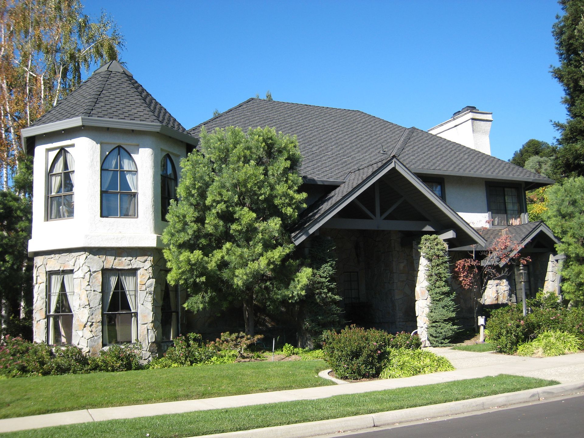 Large stone-and-stucco house with a turret, dark roof, and green lawn on a sunny day