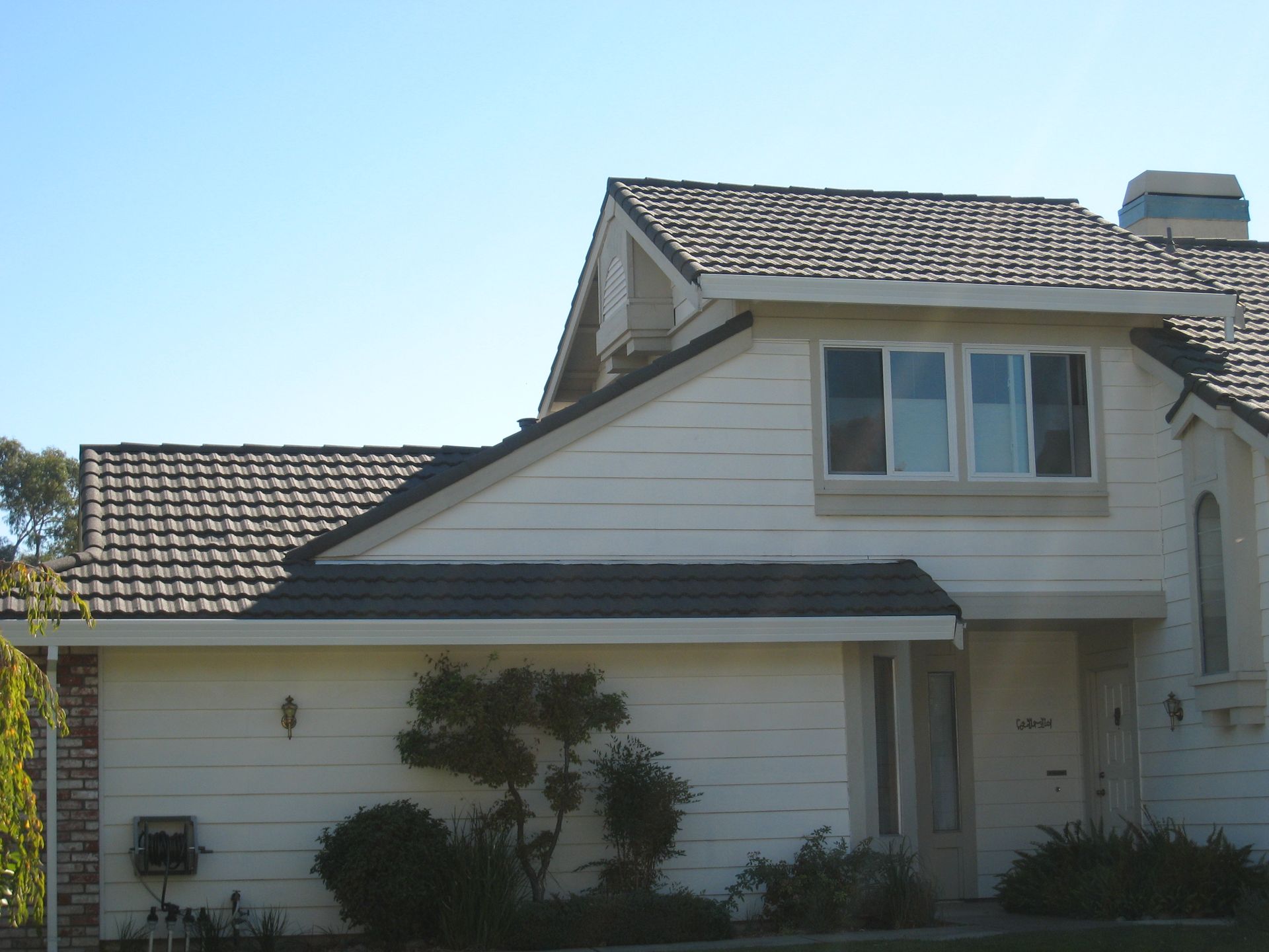White house with dark shingle roof, front porch, and shrubs under a clear blue sky