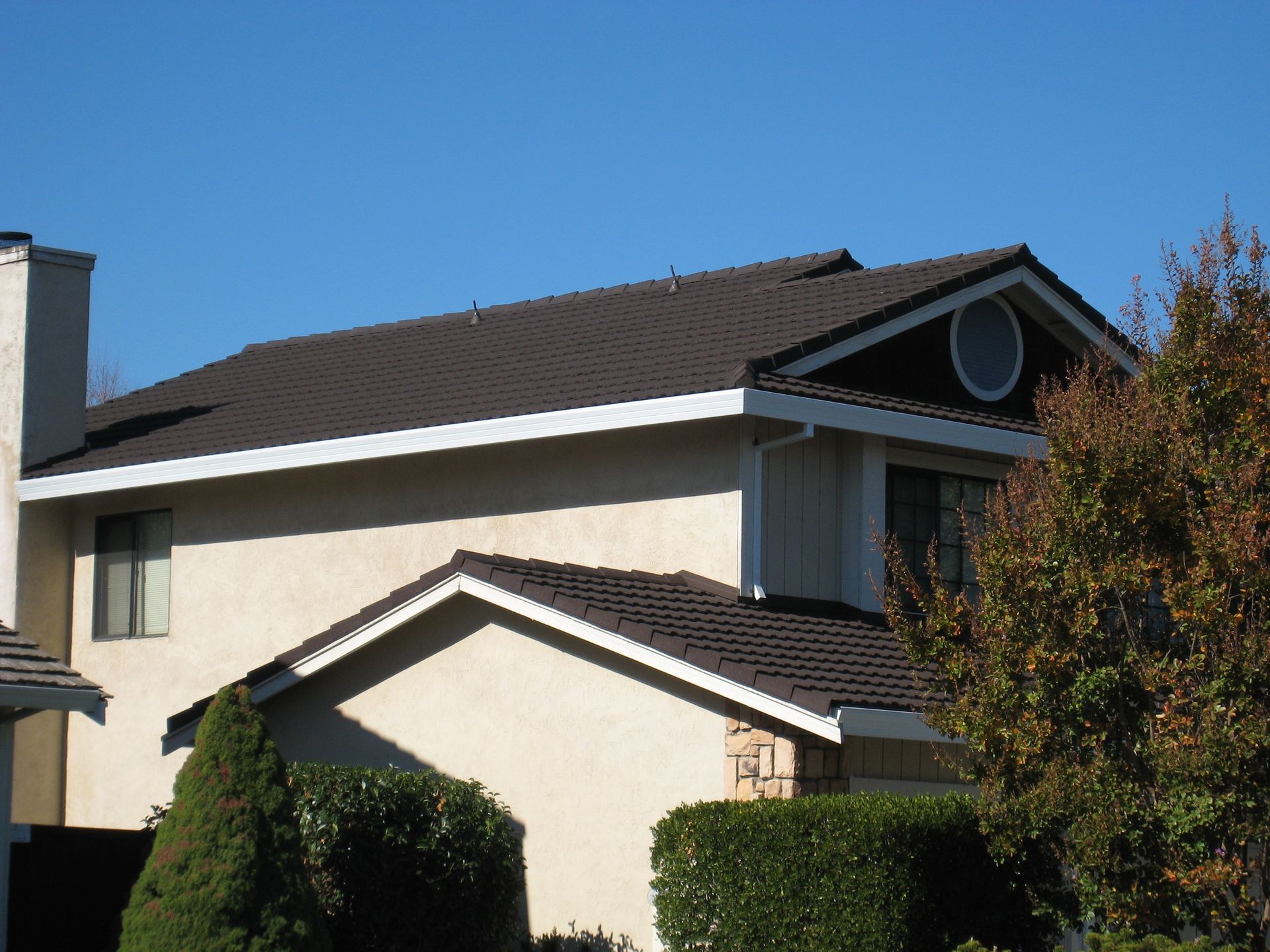 Suburban house exterior with beige walls, dark roof, and trees under a clear blue sky