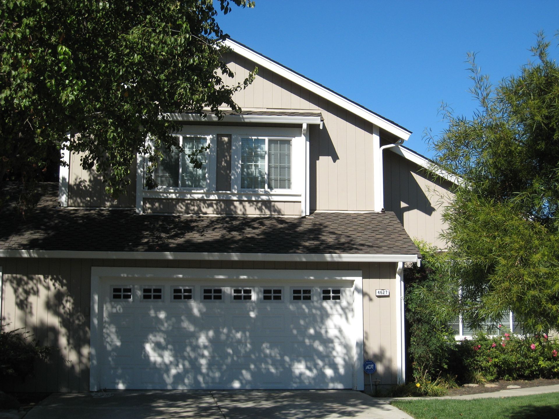 Suburban two-story house with beige siding, white garage door, and trees in front yard