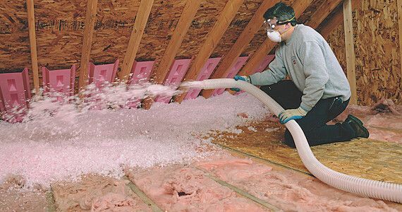 Person in an attic sprays pink foam insulation into the rafters with a hose