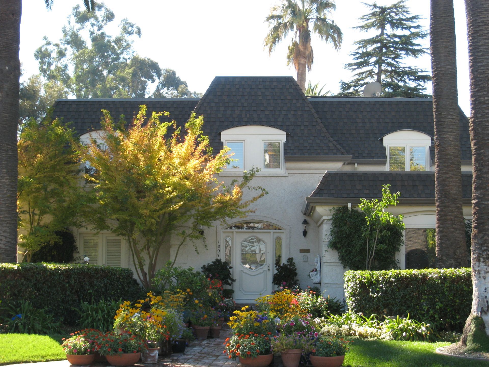 White two-story house with a dark roof, surrounded by manicured hedges, palm trees, and colorful flowers.