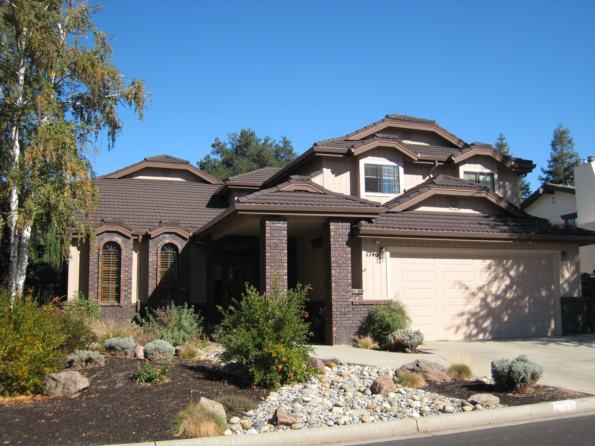 Two-story suburban house with a beige garage, brick exterior, and landscaped front yard under a clear blue sky
