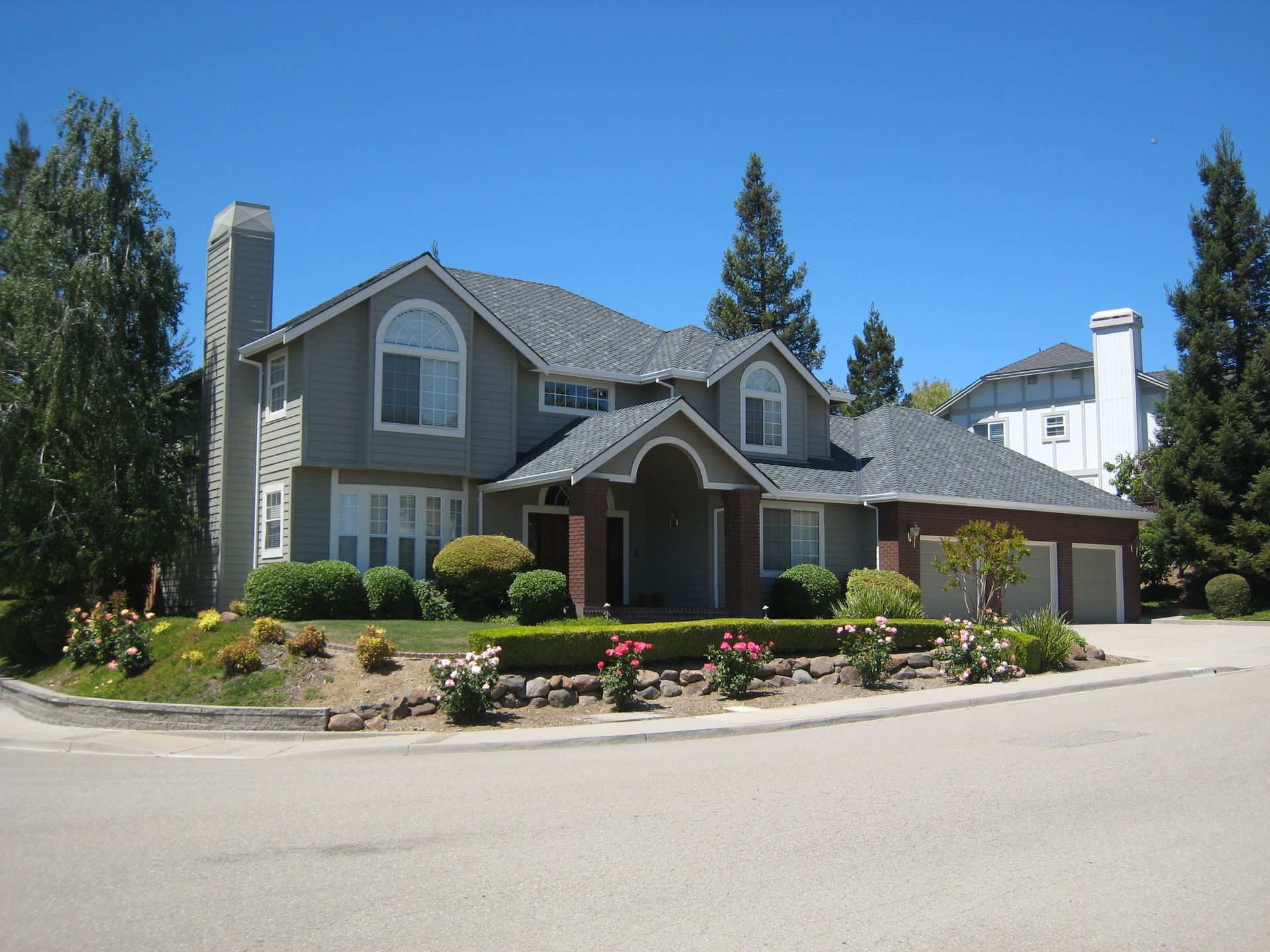 Large gray suburban house with arched entry, white trim, and landscaped front yard under a clear blue sky