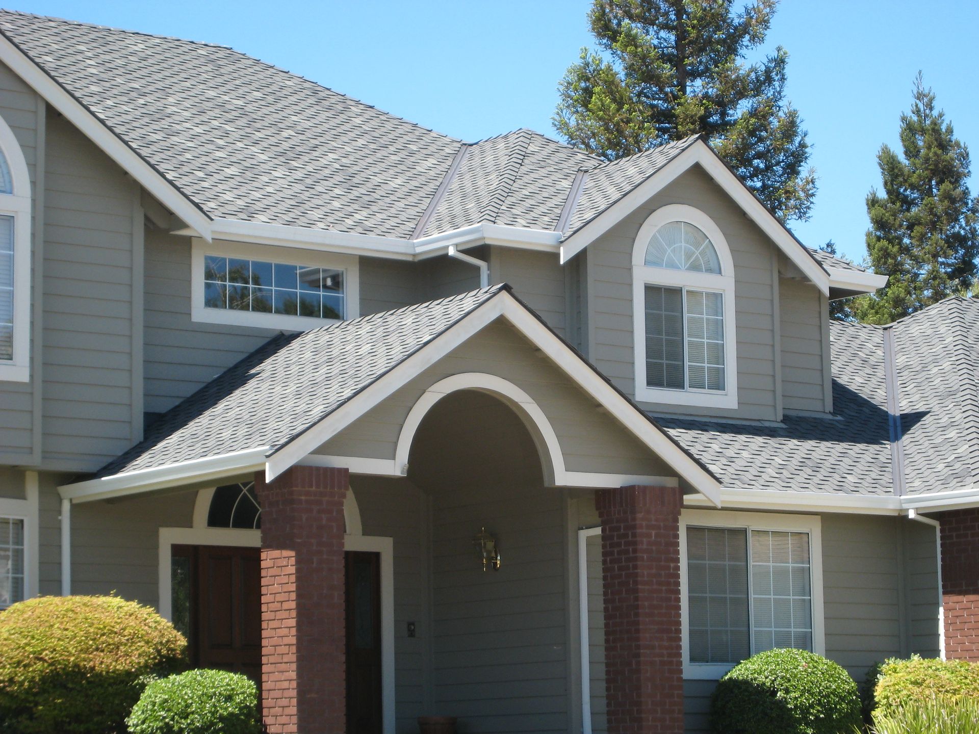 Two-story gray suburban house with brick columns and arched front porch under a blue sky