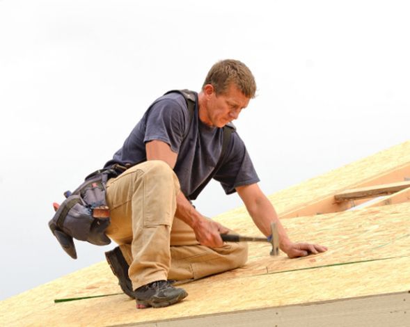 Worker kneeling on a roof, installing wooden sheathing with a power drill and hand tools