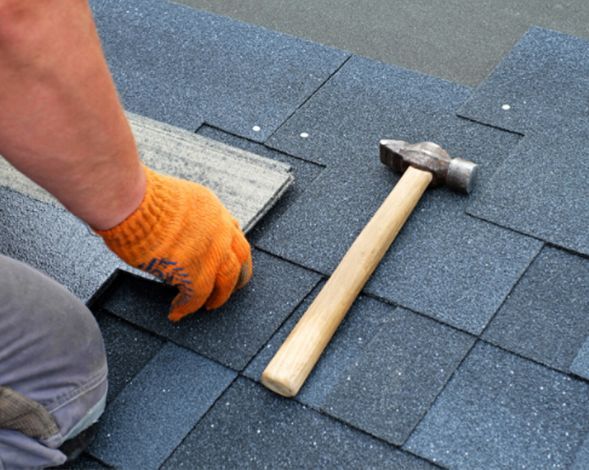 Gloved worker installing blue roof shingles with a hammer nearby