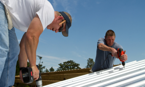 Two workers fastening white metal roofing on a sunny day, one in the foreground and one in the distance