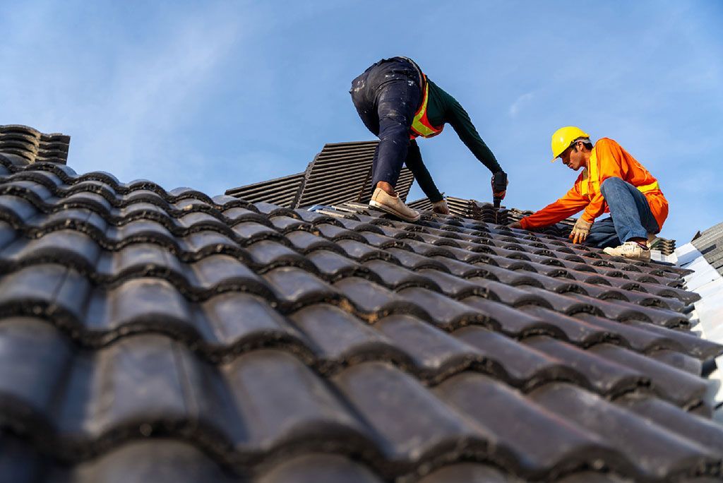 Two workers in safety gear repairing a tiled roof under a blue sky