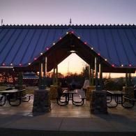 Covered outdoor picnic area at sunset with blue roof, string lights, and benches by the waterfront