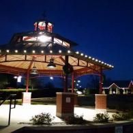 Lit pavilion at night with red roof lights and brick pillars in a landscaped park