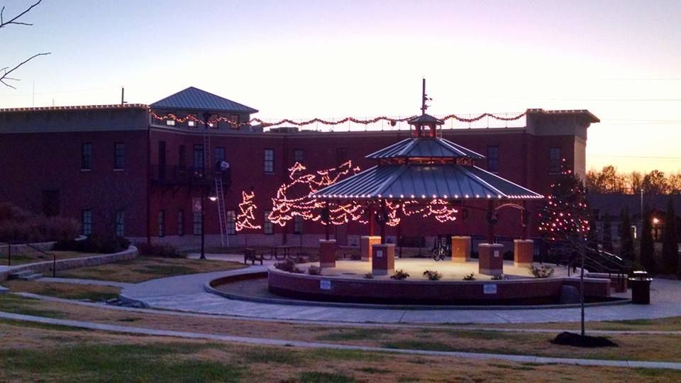 Outdoor plaza at dusk with a lit gazebo and holiday lights in front of a brick building