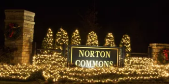 Norton Commons sign lit with warm white holiday lights at night, framed by brick pillars and trees