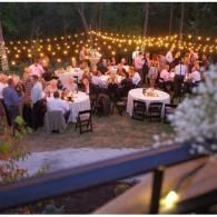 Outdoor evening wedding reception with guests seated at round tables under string lights.