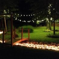 String lights over a garden path with a small bridge and glowing fire pit at night