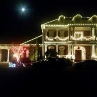 Two-story house outlined with white holiday lights at night, with a lit tree and moon overhead.