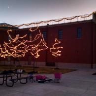 Holiday lights on a red building at dusk, with picnic tables and a cart in the foreground.