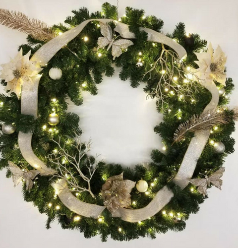 Green holiday wreath with white ribbon, flowers, and warm fairy lights on a wall