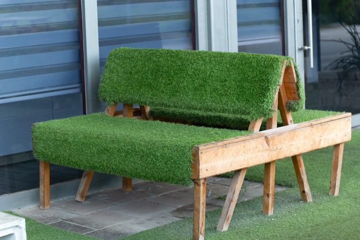 A wooden park bench covered in artificial green turf, situated outdoors near a building window.