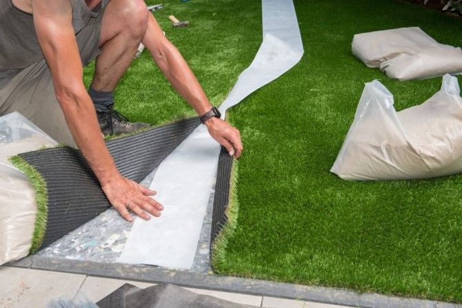 A person installs artificial grass by pressing the edge of a turf roll onto a white seam tape strip on the ground.