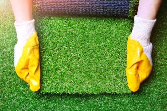 Hands wearing yellow and white work gloves are installing a rectangular piece of green artificial grass.